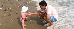 Peter crouches at the edge of the ocean, smiling and reaching out to toddler Penny standing on a beach. Waves wash around the his feet while Penny, wearing a pink patterned swimsuit and white sun hat, faces him near the shoreline.