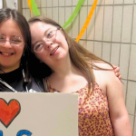 A friend and Penny standing together in a hallway, smiling and holding a handmade sign that reads “I ❤️ PALS New York.”