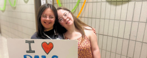 A friend and Penny standing together in a hallway, smiling and holding a handmade sign that reads “I ❤️ PALS New York.”