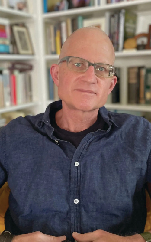 Poet Christian Wiman smiles for portrait with bookshelves blurred in the background.