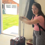 Penny who has Down syndrome gets ready to leave the house through the open front door, pushing her suitcase in front of her