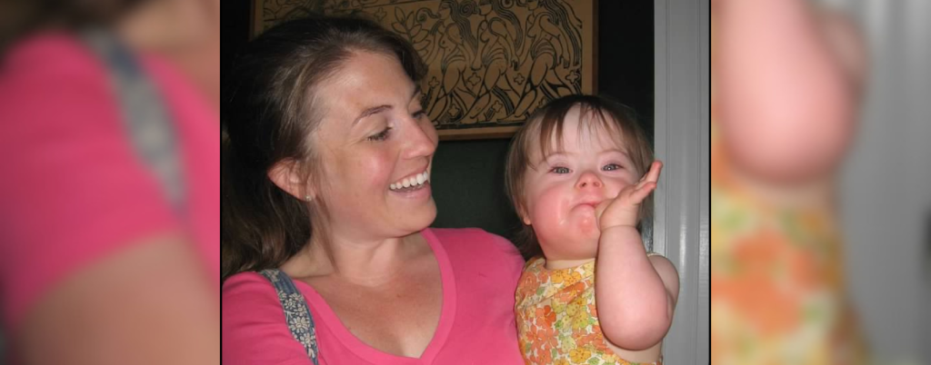 ’m holding Penny, who has Down syndrome. In the photo, she is a toddler wearing a sleeveless orange-and-yellow floral dress. She looks at the camera with a playful expression. I’m smiling at her.