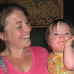 ’m holding Penny, who has Down syndrome. In the photo, she is a toddler wearing a sleeveless orange-and-yellow floral dress. She looks at the camera with a playful expression. I’m smiling at her.