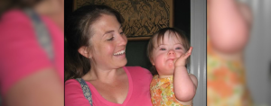 ’m holding Penny, who has Down syndrome. In the photo, she is a toddler wearing a sleeveless orange-and-yellow floral dress. She looks at the camera with a playful expression. I’m smiling at her.