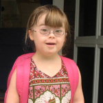 a 2010 photo of Penny wearing glasses and a pink backpack. She is smiling at the camera, dressed in a colorful patterned top