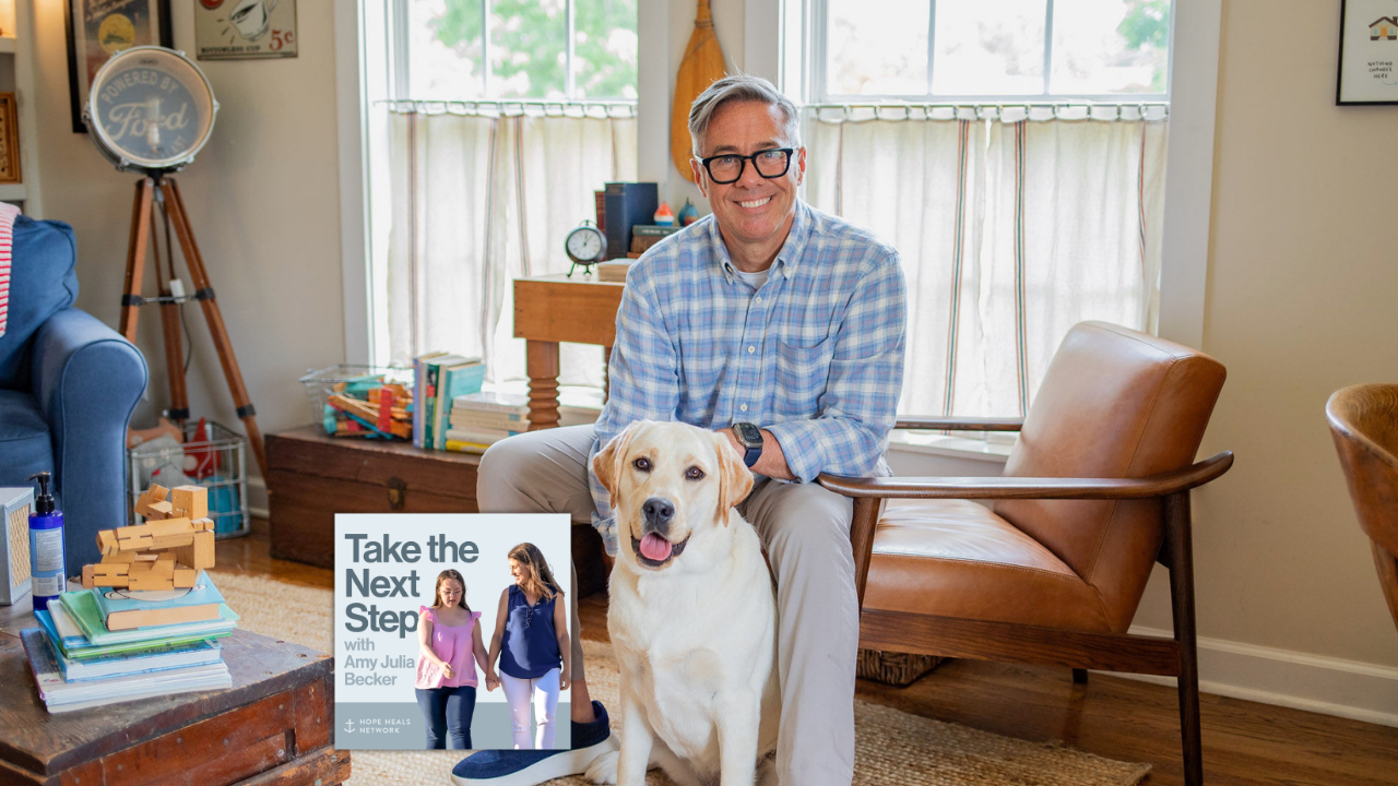 David Thomas in a blue plaid shirt sits on a chair indoors with a white-and-tan dog sitting on the floor; a living room with windows, chairs, and bookshelves is in the background. Near the middle bottom is a smaller image of the podcast cover where Penny and Amy Julia walk hand in hand, with Amy Julia looking toward Penny.