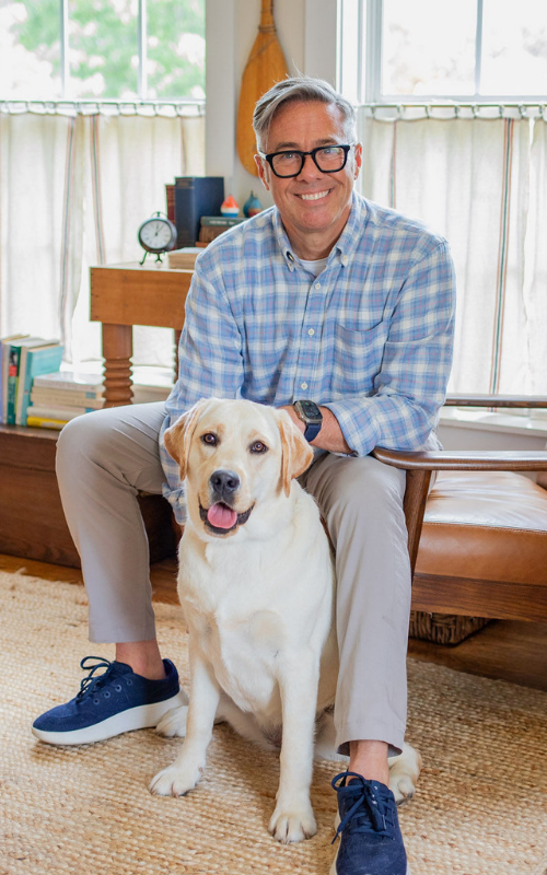 David Thomas in a blue plaid shirt sits on a chair indoors with a white-and-tan dog sitting on the floor; a living room with windows, chairs, and bookshelves is in the background.