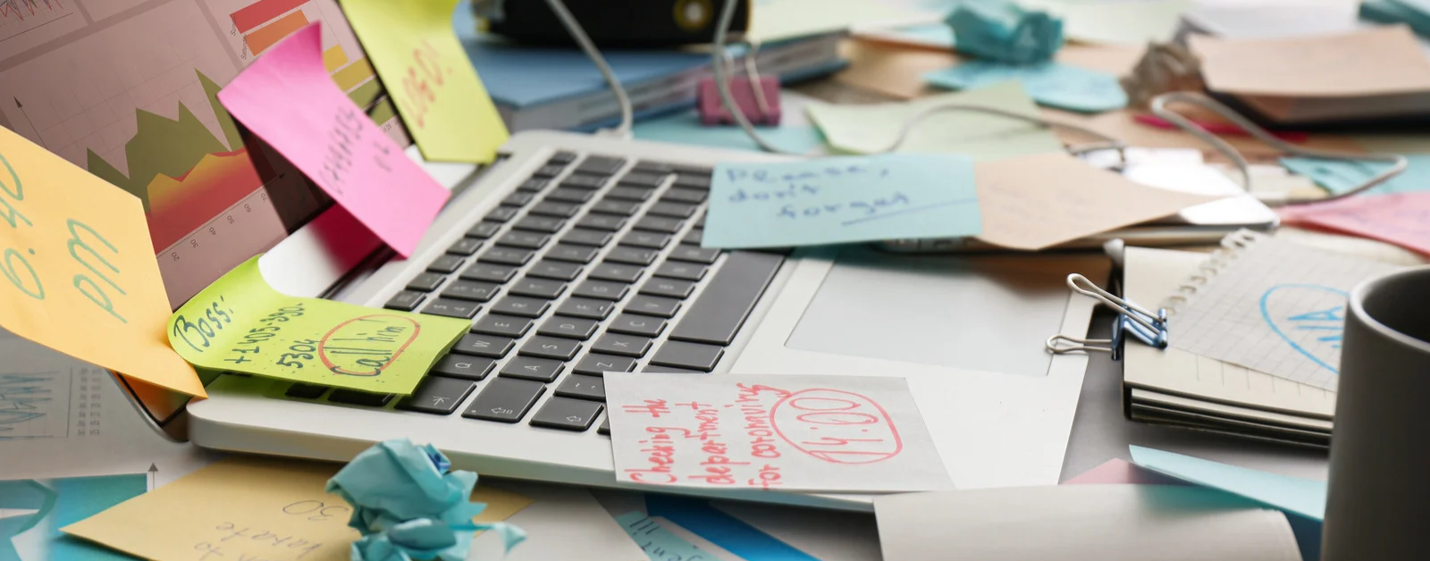 A cluttered desk with an open laptop surrounded by scattered sticky notes, papers, pens, and office supplies, with colorful notes and a blue crumpled sheet in the foreground.