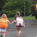 Penny and a friend wear backpacks and walk hand in hand across the street to a waiting yellow school bus