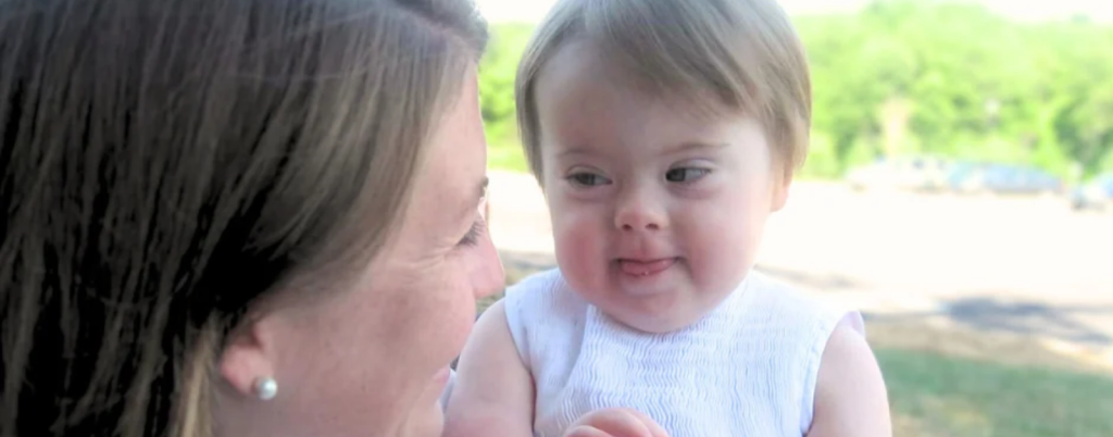 I’m holding baby Penny in my arms outdoors. She wears a white dress and looks into my eyes as I smile at her.