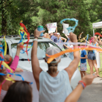 a crowd of excited people waving streamers who line a road and welcome a vehicle to Hope Heals Camp