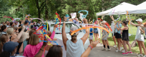a crowd of excited people waving streamers who line a road and welcome a vehicle to Hope Heals Camp