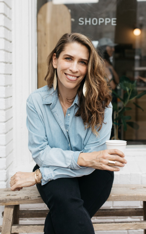 Monica DiCristina sits on a wooden bench outside a shop, wearing a light blue button-down shirt and black pants. She holds a white takeaway coffee cup and looks toward the camera.